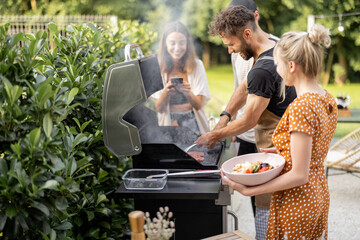 Happy young friends hanging out together, grilling vegetables and meat on a modern grill at picnic. People cooking food outdoors - obrazy, fototapety, plakaty