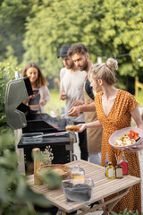 Happy young friends hanging out together, grilling vegetables and meat on a modern grill at picnic. People cooking food outdoors - obrazy, fototapety, plakaty