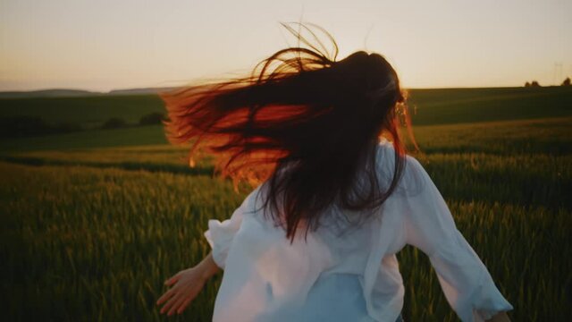 Backshot of happy delighted young woman running across rural wheat field. Excited girl in flowing white shirt enjoying sunset walk on natural meadow greenland.