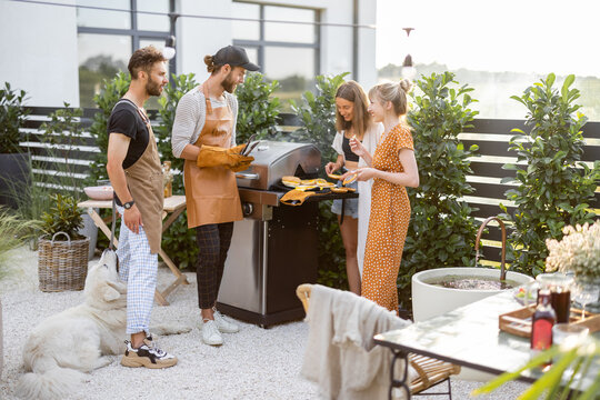 Happy young friends hanging out together, grilling food on a modern grill at beautiful backyard of a country house. People cooking food outdoors