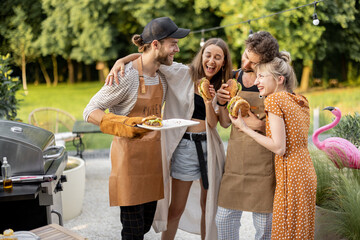 Young people enjoy yummy burgers, made on a grill at picnic, standing together and having fun. Friends cooking at backyard outdoors. American lifestyle