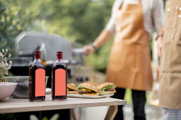 Bottles with liqueur or berry tinctures on a table with burgers, people grilling on a background, close-up on bottles with blank labels for copy paste - obrazy, fototapety, plakaty