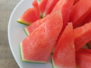A close up portrait of red pointy slices of a cut watermelon on a white plate next to an uncut green, big and round one of the fruit. A great, healthy and refreshing snack on a hot summer day.