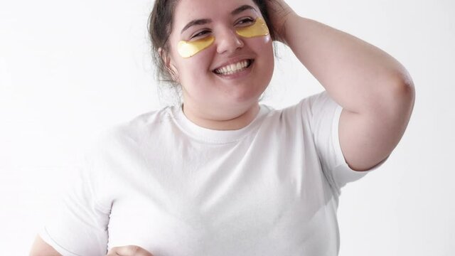Body positive. Facial skincare. Skin rejuvenation. Beauty care. Happy overweight obese woman with golden collagen hydrogel under eye patches on clean face dancing isolated on white.