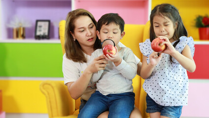 Happy asian family mother, child daughter and son sitting on sofa in colorful modern living room with smiling face while playing magnifying glasses with apples