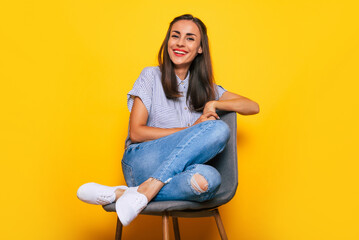 Cute smiling stylish woman is sitting on the chair and posing isolated on yellow background