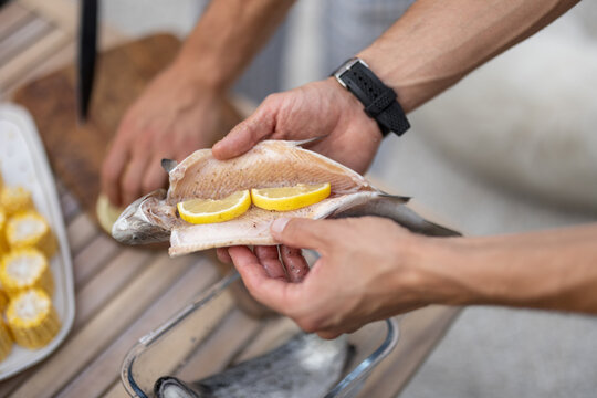Two men preparing fresh fish for grilling, seasoning it with lemon and spices at table outdoors, close-up