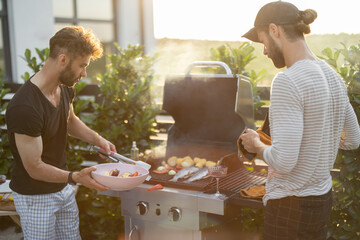 Two caucasian guys cooking vegetables and fish on a modern gas grill at backyard on a sunset....