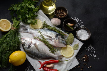 Raw fresh uncooked dorado or sea bream fish with lemon, herbs, oil, vegetables and spices on on white paper over black background