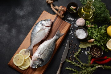 Raw fresh uncooked dorado or sea bream fish with lemon, herbs, oil, vegetables and spices on rustic wooden board over black background