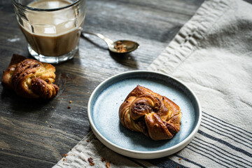 Cinnamon yeast knot bun on blue plate and glass of coffee with milk on dark wooden table.