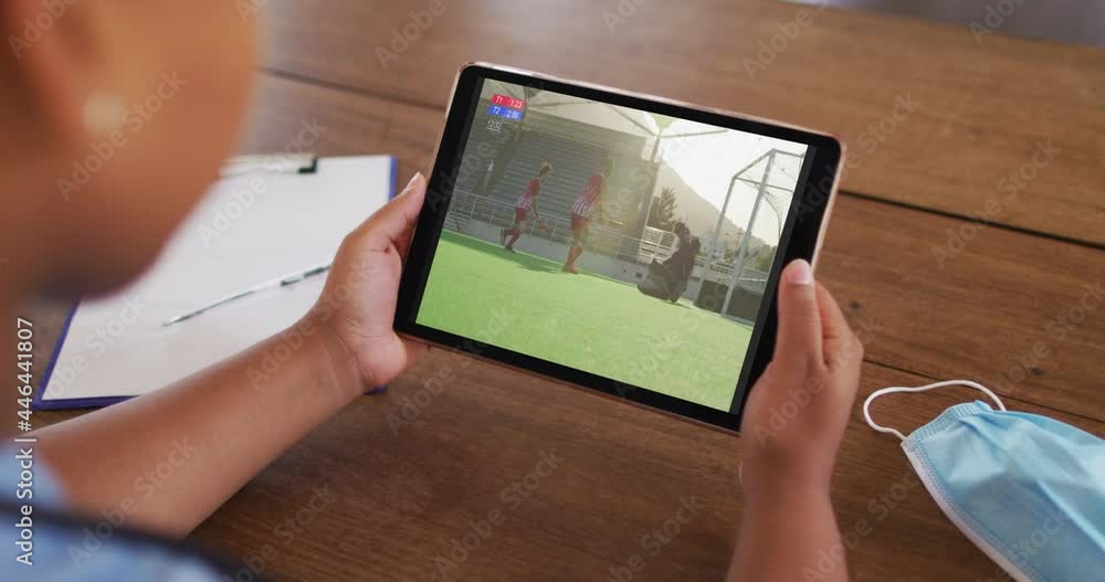 Wall mural Composite of woman sitting at table with face mask, watching hockey match on tablet