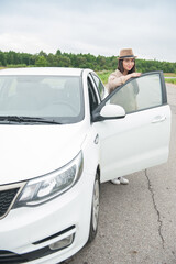 Beautiful happy young woman with hat standing near the white car on the road