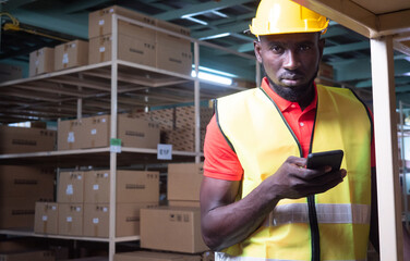 African engineer or technician worker wearing safety helmet and vest use a cell phone in modern automatic warehouse. Warehousing, machinery concept. Logistics in stock.