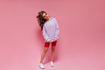 Long-haired girl in cycling shorts and purple oversized hoodie poses on pink background. Portrait...
