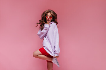 Brunette curly woman in red sunglasses, purple hoodie and cycling shorts jumps on isolated. Cool girl looks surprised on pink background.