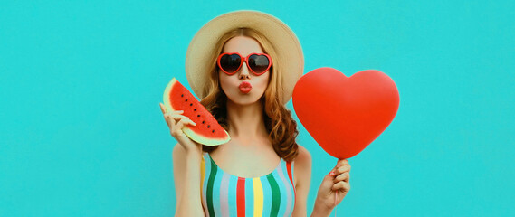 Portrait of beautiful young woman with slice of fresh watermelon and big red heart shaped balloon blowing her lips wearing a summer straw hat, sunglasses on blue background