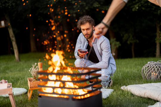 Two guys light a fire in a barbecue preparing for a picnic on a green lawn near the forest in the evening