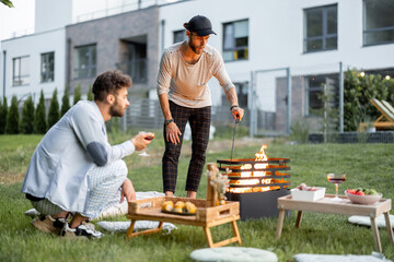 Two male friends have a picnic, making fire at barbeque on the green lawn at backyard of the...