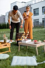 Young couple have a picnic, making fire at barbeque and drink wine on the green lawn at backyard of...