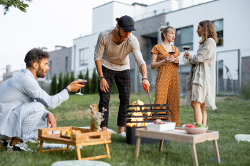 Friends have a picnic, making fire at barbeque and talking on the green lawn at backyard of the country houses on the evening