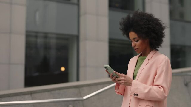 Beautiful young businesswoman wearing formal suit and using modern smart phone while walking at break in the city. Professional female employer typing text message on cellphone outside.