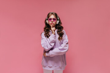 Thoughtful woman in purple hoodie and white pants poses on isolated. Brunette girl listening to music in headphones on pink background.