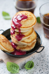 Serving pan with mini pancakes and jam, vertical shot on a beige granite surface with coffee glasses in the background, close-up