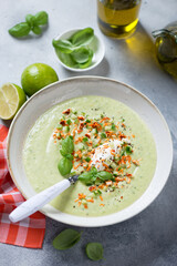 Plate of cold cucumber gazpacho soup topped with cheese and green basil, vertical shot on a light-blue stone surface