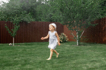 A happy little girl in a dress runs along the garden lawn and plays with a lawn sprinkler.