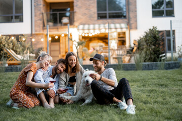 Young friends having fun playing with a dog and drinking wine, sitting on the green lawn at backyard of the country house in the evening