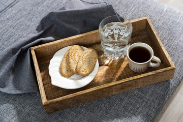 A white cup of coffee, a glass of water, a long cupronickel spoon with a twisted handle and a saucer with lemon for breakfast on a wooden tray, a rag napkin on a gray sofa.