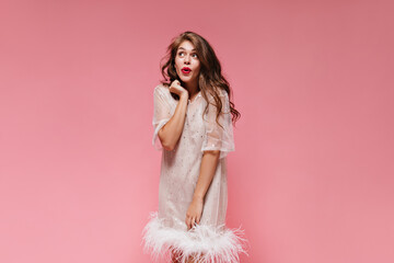 Portrait of curly brunette woman in white dress posing in good mood on pink background. Charming girl looks dreamily on isolated.