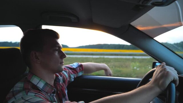 Profile of handsome man in shirt driving car through countryside. Young guy riding on auto with his hand out of window enjoying road trip. Beautiful view at background. Slow motion Close up