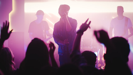 Rock Band with Guitarists and Drummer Performing at a Concert in a Night Club. Front Row Crowd is Partying. Silhouettes of Fans Raise Hands in Front of Bright Colorful Strobing Lights on Stage. 