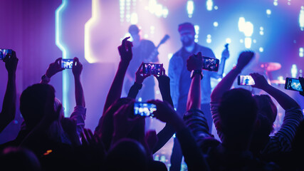 Rock Band with Guitarists and Drummer Performing at a Concert in a Night Club. Front Row Crowd is Record Video on Their Mobile Phones. Party in Front of Bright Colorful Strobing Lights on Stage. 