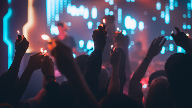 Rock Band Performing a Slow Song at a Concert in a Night Club. Front Row Crowd is Holding Lighters. Silhouettes of Fans Raise Hands in Front of Bright Colorful Strobing Lights on Stage.