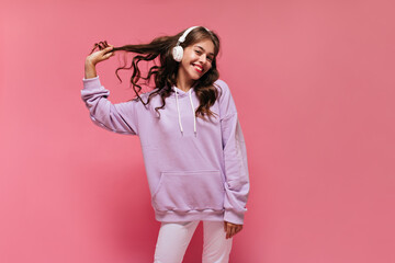 Cheerful girl in purple hoodie and white pants smiles sincerely. Happy curly woman listens to music...