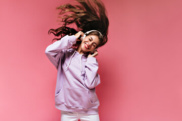 Cheerful happy girl in purple hoodie plays hair and listens to music in headphones. Curly woman dances and smiles on isolated pink background.