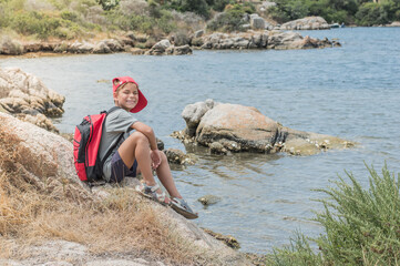 A happy traveler boy with backpack is sitting on rocks near the sea
