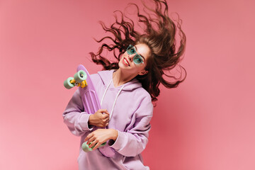 Curly brunette woman in purple oversized hoodie plays hair. Cool teen girl in green sunglasses smiles and holds colorful longboard on isolated.