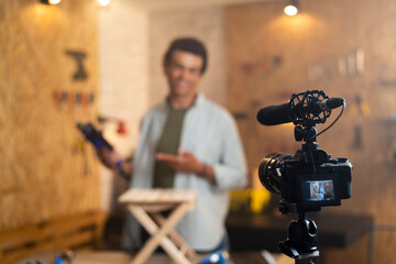 DIY carpenter filming a blog about drill with focus on camera and a carpenter blurred in the back