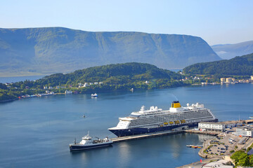 Cruise ship with blue hull and yellow funnel docked in the harbour in the Port of Alesund, Norway. Beautiful fjord landscape with mountain in the background and town in the forground.