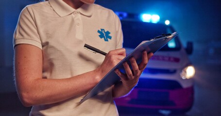 Close up of female paramedic writing with pen in hand in folder with medical cards and documents. Woman medic filling in medical form at night outdoor. Emergency situation. Call 911, paramedic
