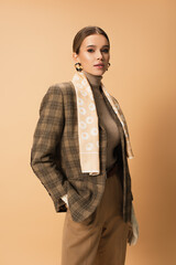 stylish woman in blazer, pants and white gloves posing with hand in pocket on beige