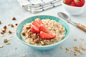 Oatmeal porridge with strawberry slices, nuts almonds and honey in blue bowl on grey table. Healthy eating, dieting, vegetarian food concept. Place for text