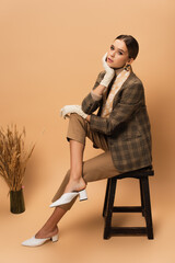 elegant woman in checkered blazer, pants, white shoes and gloves sitting near vase with spikelets...