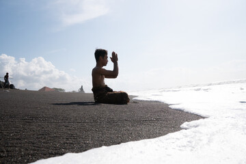 A Balinese Hindu man praying on the beach on a hot sunny day. The sand is black and the waves are foamy white