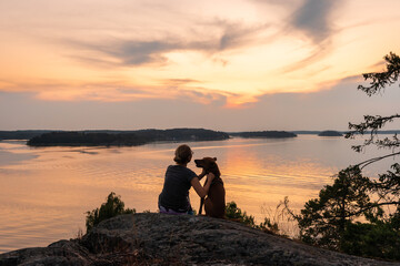 Beautiful sunset on the Baltic Sea. Bright golden orange pink tones of the sunset sky. Islands of archipelago. Young woman with her dog sitting on top of high stone cliff and watches the setting sun.