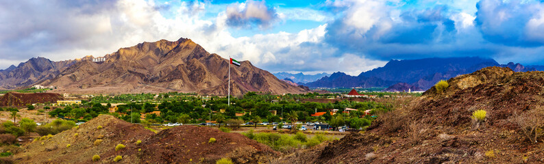 Hatta city welcoming sign written with large letters placed in Hajar mountains and UAE flag flying high in Hatta enclave of Dubai in the United Arab Emirates. - obrazy, fototapety, plakaty